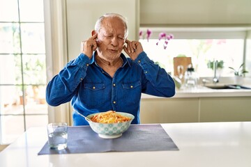 Senior man with grey hair eating pasta spaghetti at home covering ears with fingers with annoyed...