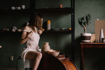 talented sculptor girl in thin light negligee sculpts a jug of clay in pottery workshop near large window. Concept art of sculpture
