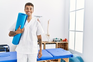 Young hispanic man working as physiotherapist holding yoga mat at physiotherapy room