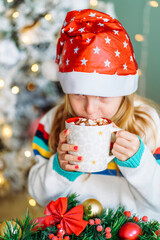 Little smiling girl Santa helper drink hot chocolate with marshmallows from christmas mug with with christmas wreath on background