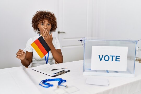 Young african american woman at political campaign election holding germany flag covering mouth with hand, shocked and afraid for mistake. surprised expression