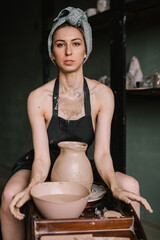 young sculptor, a girl with bandage on her head and in black apron, sculpts a clay product in pottery workshop near large window. Concept hobby sculpture