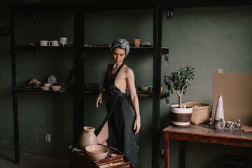 Obraz na płótnie Canvas beautiful sculptor girl with bandage on her head and a black apron on her naked body is standing in pottery workshop near large window. concept is beauty and art.