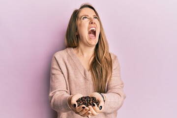 Young blonde woman holding coffee beans with hands angry and mad screaming frustrated and furious,...