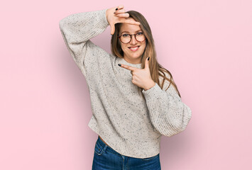Young blonde woman wearing casual sweater and glasses smiling making frame with hands and fingers with happy face. creativity and photography concept.