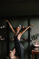 beautiful sculptor girl with bandage on her head and a black apron on her naked body is standing in pottery workshop near large window. concept is beauty and art.