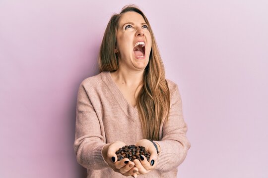 Young blonde woman holding coffee beans with hands angry and mad screaming frustrated and furious, shouting with anger looking up.