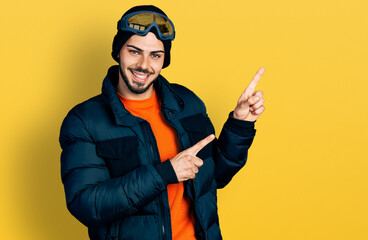 Young hispanic man with beard wearing snow wear and sky glasses smiling and looking at the camera pointing with two hands and fingers to the side.