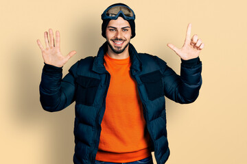Young hispanic man with beard wearing snow wear and sky glasses showing and pointing up with fingers number seven while smiling confident and happy.