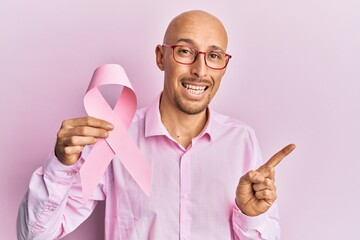 Bald man with beard holding pink cancer ribbon smiling happy pointing with hand and finger to the side