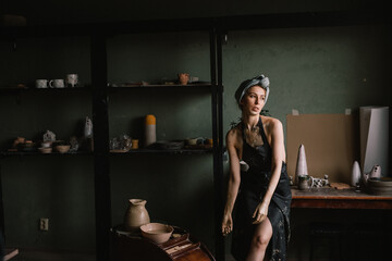 beautiful sculptor girl with bandage on her head and a black apron on her naked body is standing in pottery workshop near large window. concept is beauty and art.