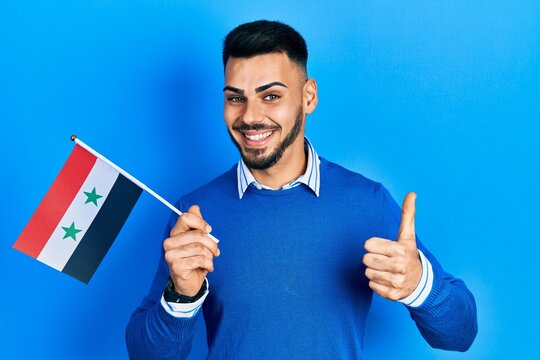 Young hispanic man with beard holding syria flag smiling happy and positive, thumb up doing excellent and approval sign