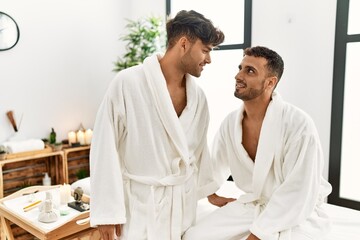 Two hispanic men couple wearing bathrobe sitting on massage table at beauty center - obrazy, fototapety, plakaty