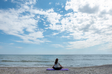Fototapeta na wymiar Peaceful beauty. Meditating woman. Sea harmony. Unrecognizable lady practicing yoga in lotus pose on azure water coast in sunny daylight.