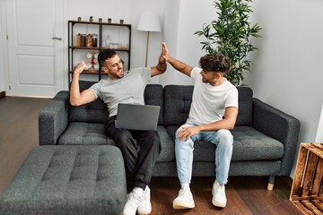 Two hispanic men couple using laptop high five with hands raised up at home