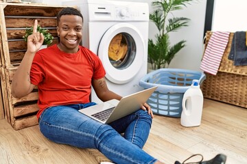 Young african man doing laundry and using computer showing and pointing up with finger number one while smiling confident and happy.
