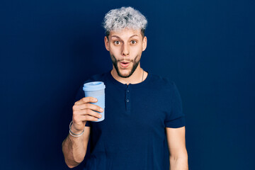 Young hispanic man with modern dyed hair drinking a take away cup of coffee scared and amazed with open mouth for surprise, disbelief face