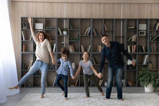 Portrait of overjoyed family with two kids holding hands, jumping at home, excited happy mother and father with little daughter and son engaged in funny activity in living room, dancing and laughing