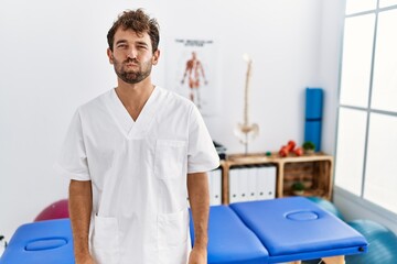 Young handsome physiotherapist man working at pain recovery clinic puffing cheeks with funny face. mouth inflated with air, crazy expression. - obrazy, fototapety, plakaty