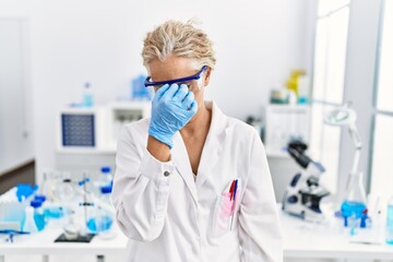 Middle age blonde woman working at scientist laboratory tired rubbing nose and eyes feeling fatigue...
