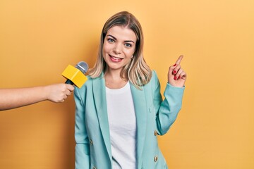 Beautiful caucasian woman being interviewed by reporter holding microphone smiling happy pointing...