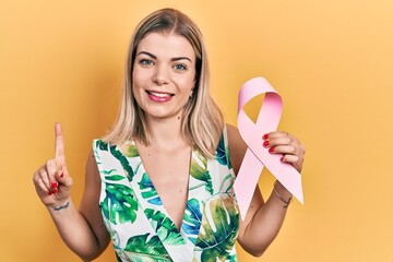 Beautiful caucasian woman holding pink cancer ribbon smiling with an idea or question pointing finger with happy face, number one