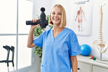 Young blonde woman wearing physiotherapist uniform holding massage pistol at rehab clinic