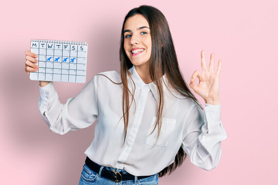 Young brunette teenager holding travel calendar doing ok sign with fingers, smiling friendly gesturing excellent symbol