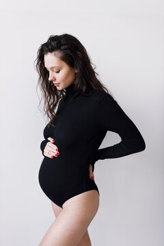 A young beautiful pregnant woman in a black bodysuit, on a white wall. The concept of a fashionable image for pregnant women.