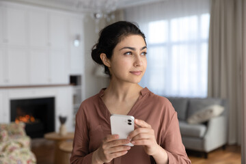 Attractive pensive Indian woman standing in living room holds smartphone looking into distance....