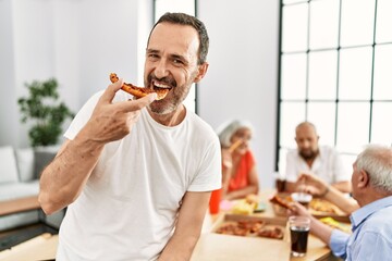 Group of middle age people smiling happy eating italian pizza sitting on the table at home