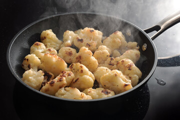 steaming roasted cauliflower florets in a black frying pan, cooking vegetables for a healthy...