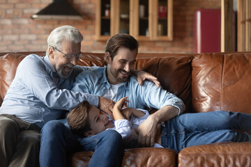 Funny playtime. Joyful elderly grandfather millennial dad tickle giggling tween kid boy on couch enjoy cute active game. Overjoyed three generation men family laugh aloud relax play have fun together © fizkes