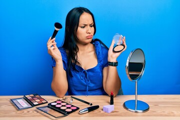 Beautiful hispanic woman with nose piercing holding makeup powder and brush skeptic and nervous,...