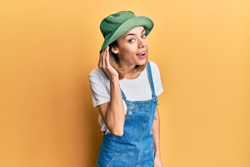 Young caucasian blonde woman wearing denim jumpsuit and hat with 90s style smiling with hand over...