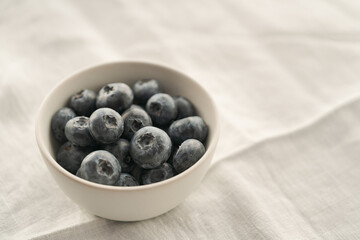 Organic ripe blueberries in white ceramic bowl on white linen cloth