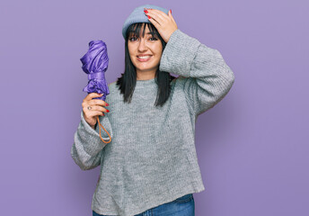 Young hispanic woman holding closed purple umbrella stressed and frustrated with hand on head, surprised and angry face