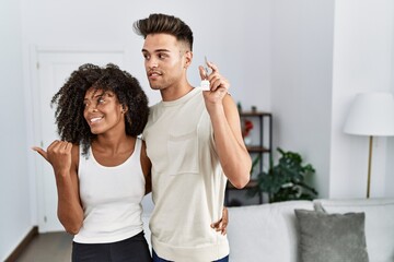 Young interracial couple holding keys of new home pointing thumb up to the side smiling happy with open mouth