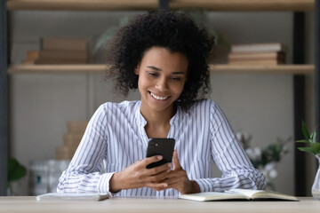 Head shot smiling African American woman using smartphone, chatting online, happy young female looking at phone screen, watching video in social network, browsing surfing internet, sitting at desk