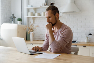 Thoughtful young hipster man looking at computer screen, considering problem solution or pondering tasks, creating new ideas for project, doing remote freelance work, sitting at table in kitchen.