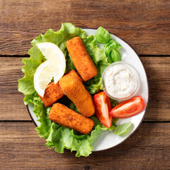 Crumbed fish sticks served with lemon, lettuce leaves and tartar sauce, healthy eating for kids. Wooden background, top view