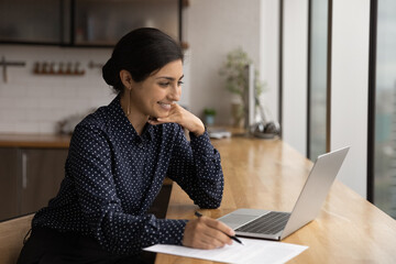 Happy young Indian woman looking at laptop screen, holding video web camera call distant conversation with colleagues or listening educational lecture, writing notes studying on online courses.