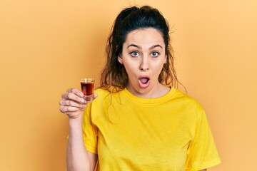 Young hispanic woman drinking whiskey shot scared and amazed with open mouth for surprise,...