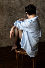 Young woman in stockings and blue oversize shirt sits on chair legs tucked up and facing the wall