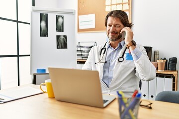 Middle age caucasian man wearing doctor uniform talking on the smartphone al clinic