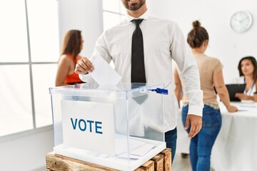 Young voter man smiling happy putting vote in voting box standing by ballot at electoral center.