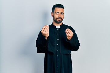 Handsome hispanic man with beard wearing catholic priest robe doing money gesture with hands,...