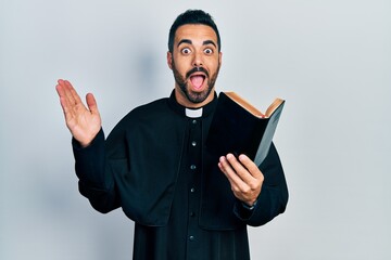 Handsome hispanic priest man with beard holding bible and christian cross celebrating crazy and...