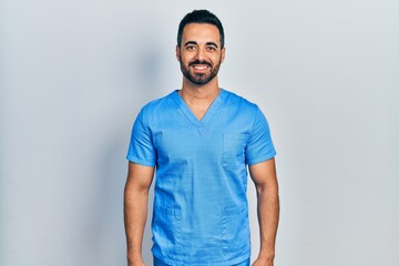 Handsome hispanic man with beard wearing blue male nurse uniform with a happy and cool smile on...
