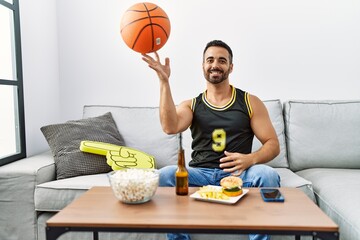Young hispanic man supporting basketball game playing with ball at home